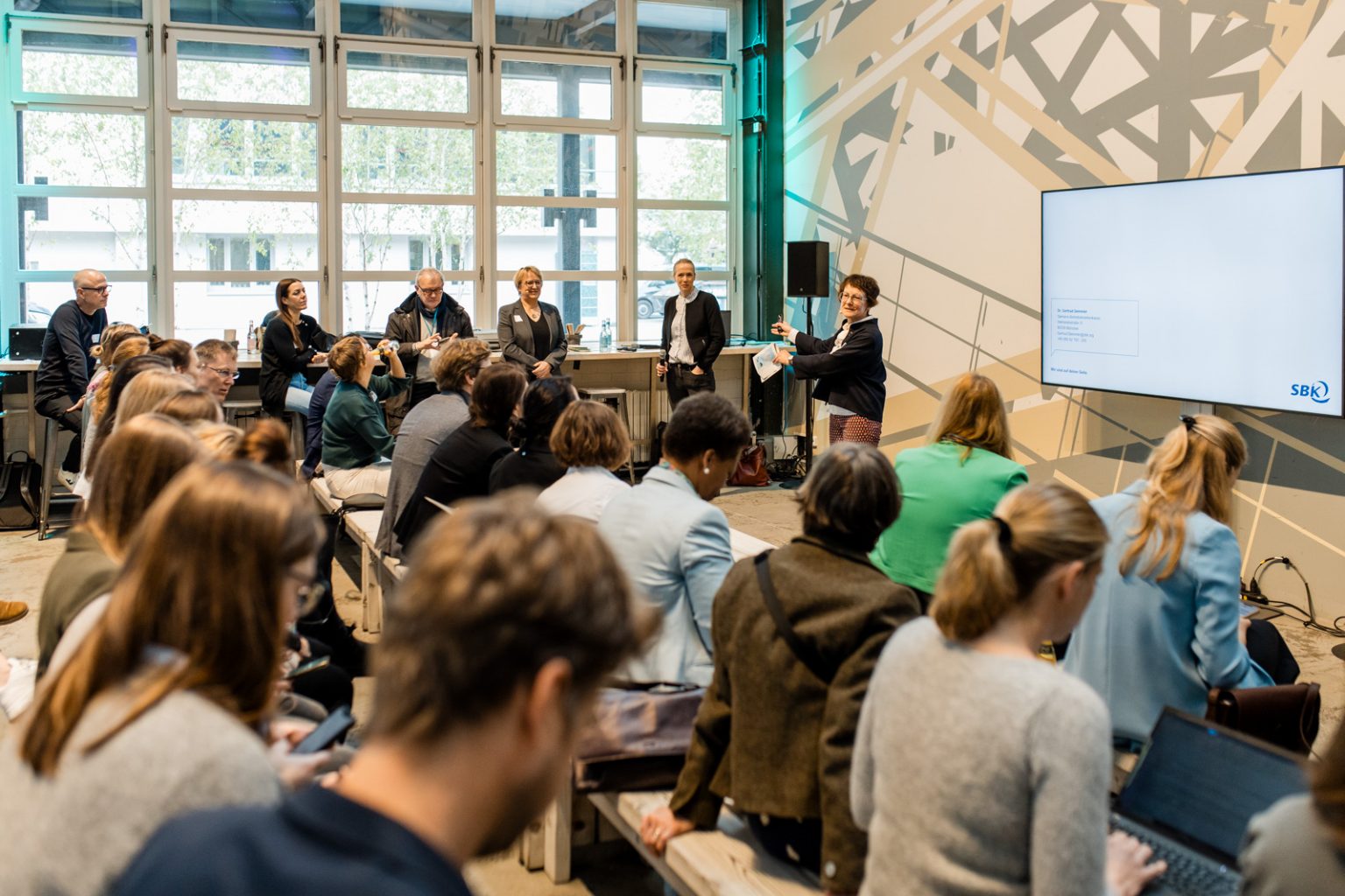 Eine Frau präsentiert etwas auf einer kleinen Bühne in der Werkstatt, Menschen schauen ihr von Sitzreihen aus zu