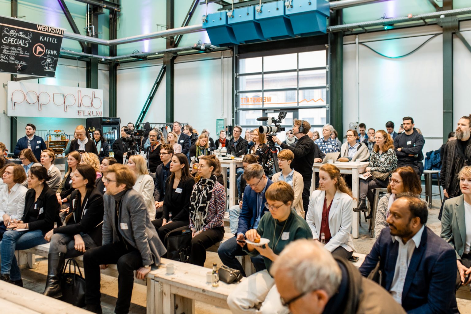 Talk im Foyer der Werkstatt, die Gäste sitzen auf Bänken, ein Kameramann nimmt auf