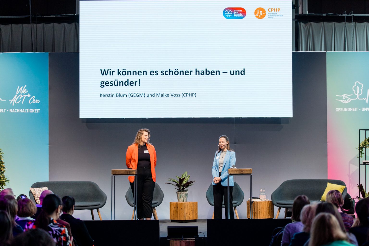 Nahaufnahme von einer Bühne mit 2 Moderatorinnen, eine große Leinwand hängt über ihren Köpfen