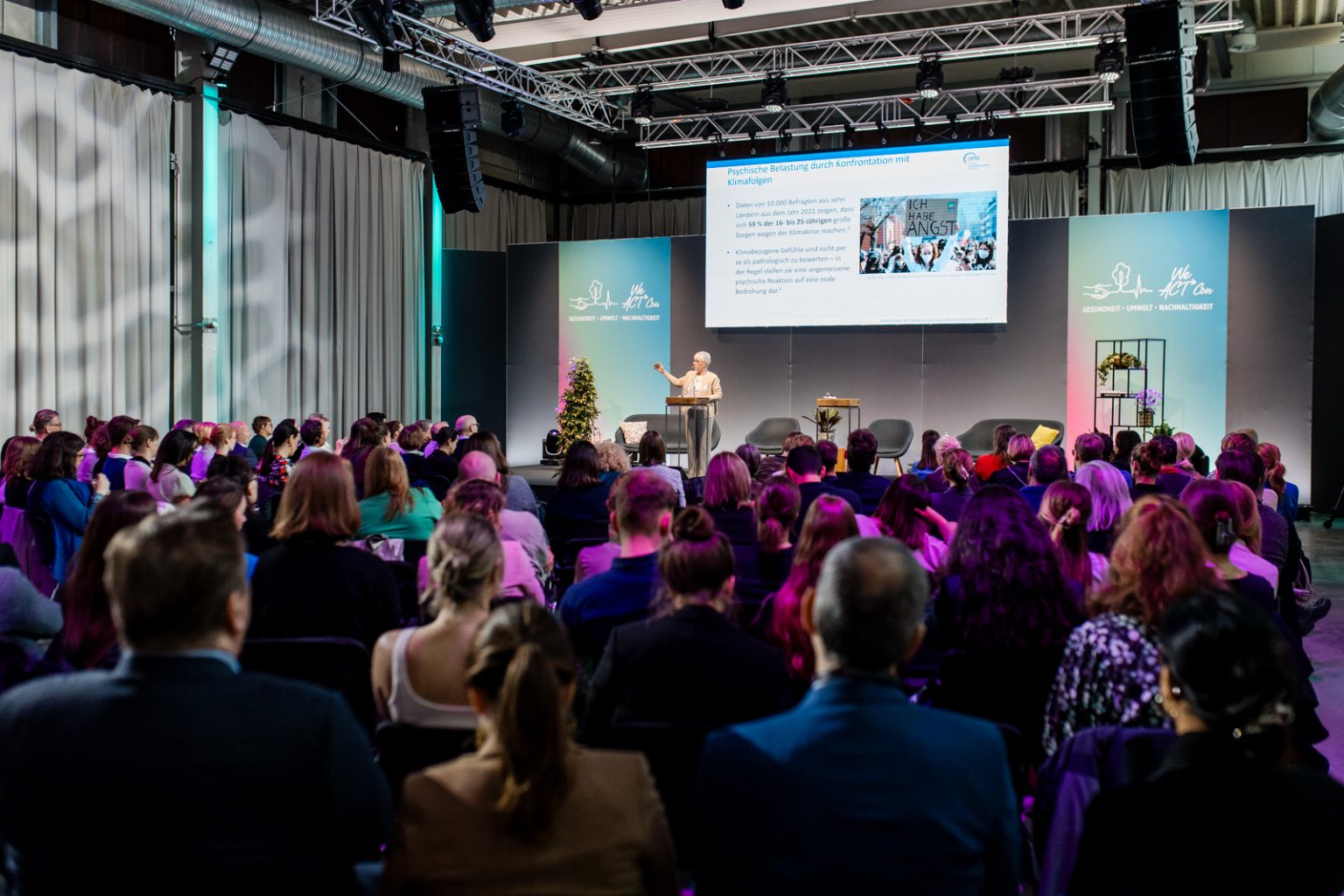 Eine Konferenz in der W26, Menschen schauen auf eine Bühne mit einer Rednerin