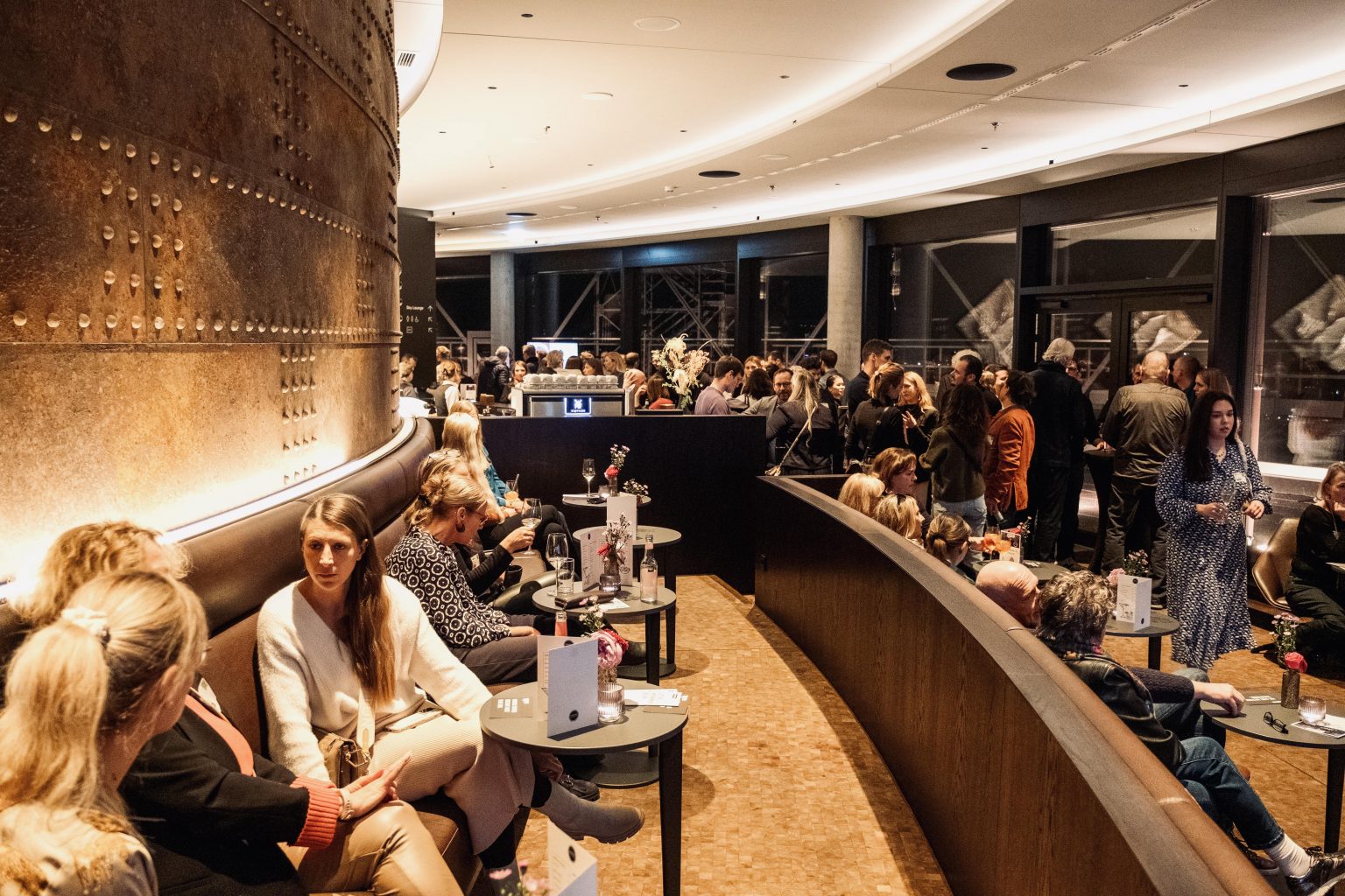 Gäste in der Gasometer Sky Bar bei geselligen Gesprächen, das Licht ist warm, auf Tischen stehen Gläser, eine Bar ist im Hintergrund zu sehen.