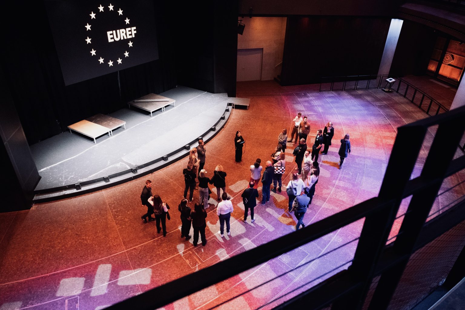 Eine Gruppe von Menschen erhält eine Führung im Gasometer Forum, der Raum ist von oben fotografiert, eine Menschentraube steht vor der Bühne.