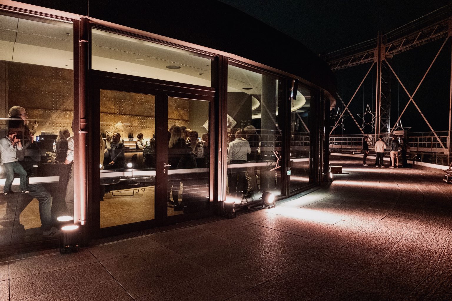 Die Dachterrasse des Gasometers am Abend: Weißes Licht beleuchtet den Außenbereich, durch die Glasscheibe blickt man nach innen auf eine Veranstaltung und die Gäste