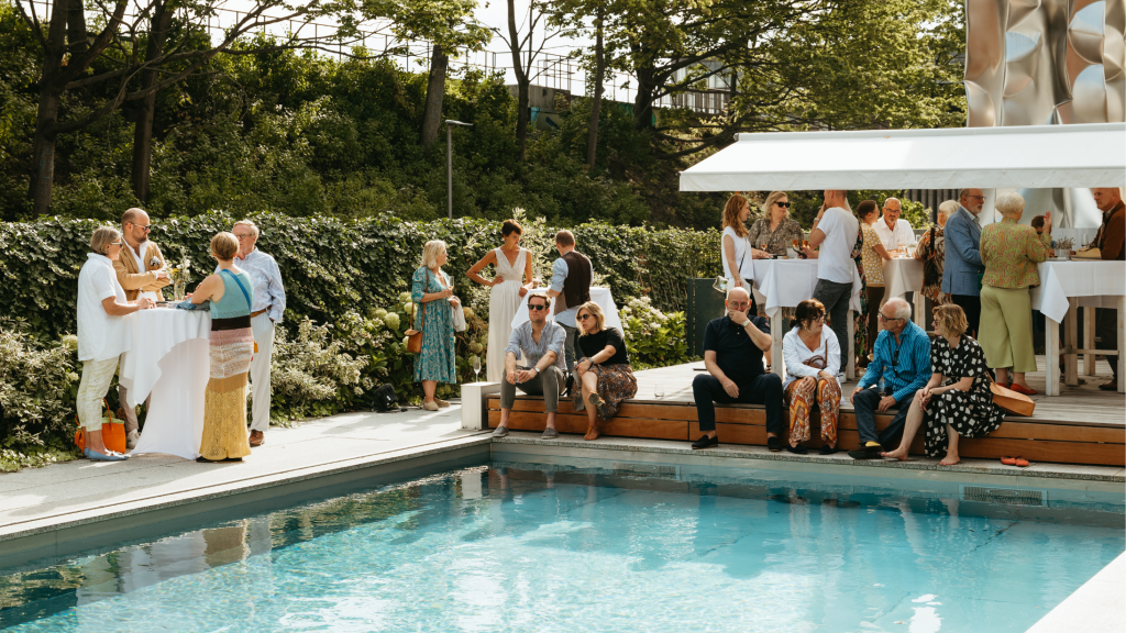 Bei einer Sommerveranstaltung sitzen Gäste an und um einen Pool herum, die Sonne scheint, die Stimmung ist ausgelassen.