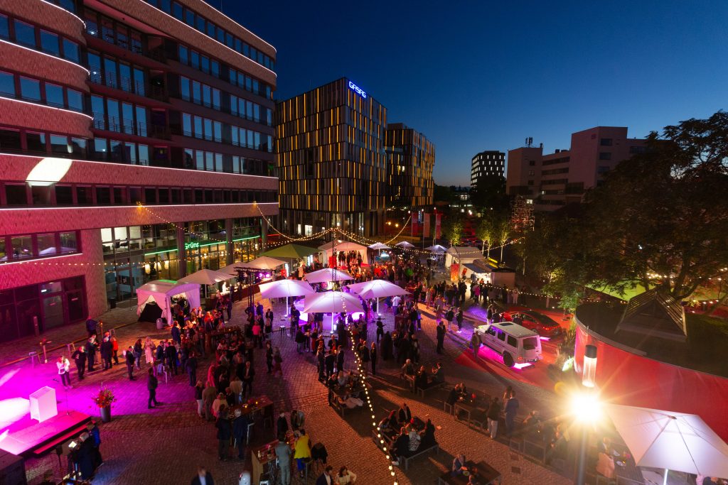 Blick auf den EUREF-Plaza bei Abenddämmerung: der Platz leuchtet in pink und sommerlichen Lichterketten, Menschen stehen und sitzen zusammen, es ist ein ausgelassenes Fest