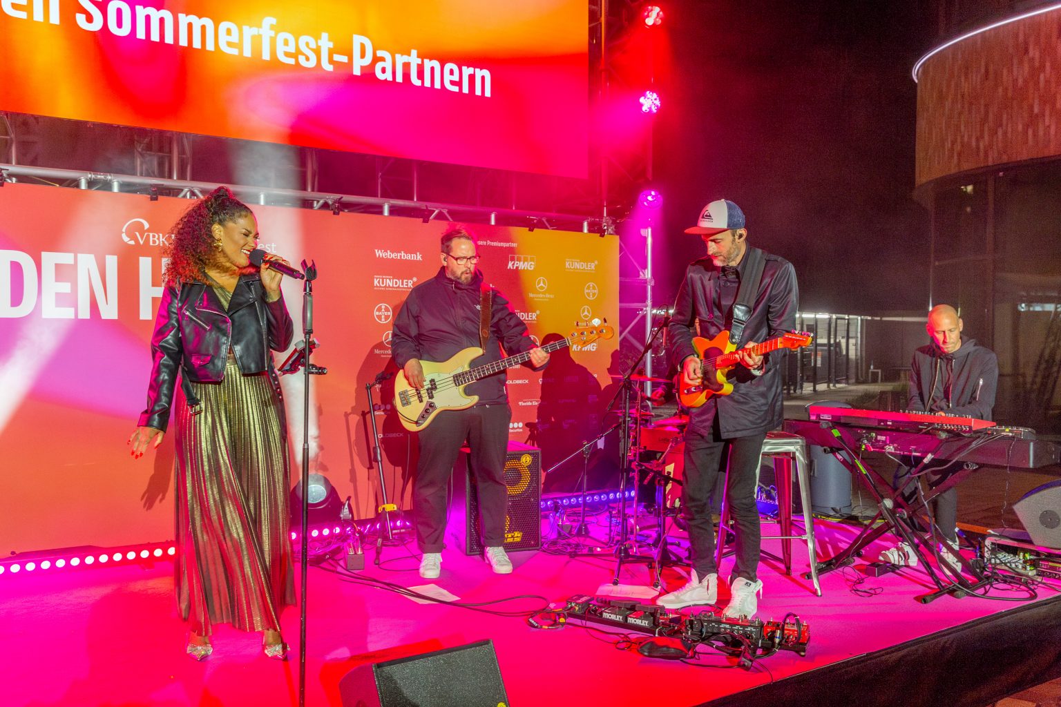 Eine Band spielt auf der Bühne unterhalb der LED Wand, alles leuchtet in pink und orange