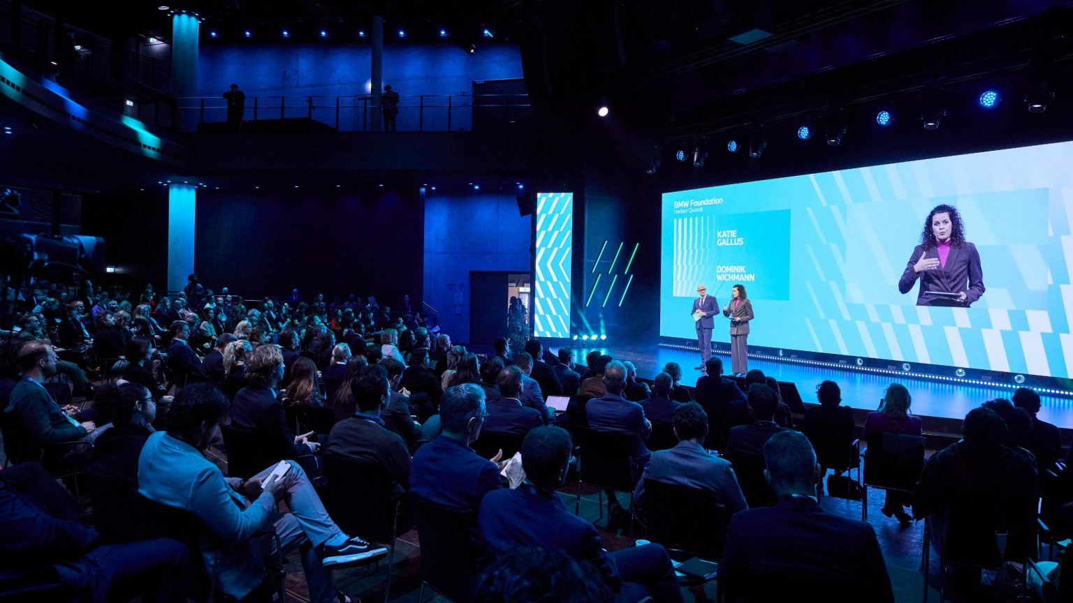 Zuschauer schauen auf die Bühne im Gasometer, zwei Moderatoren sprechen zum Publikum