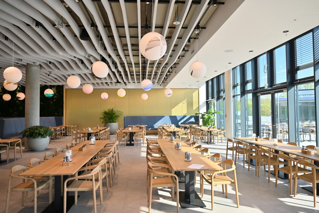 Heller Raum mit Fensterfront und langen Holztischen, und Stühlen, der als Gastronomie oder Eventlocation genutzt wird.
