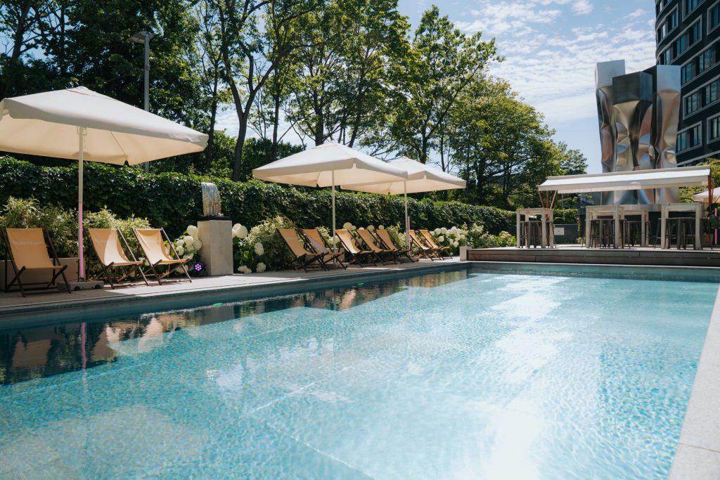 Der EUREF-Pool auf dem Berliner Campus, mit gelbel Liegestühlen umrandet, 3 große Sonnenschirme spenden Schatten, im Hintergrund sind Bäume