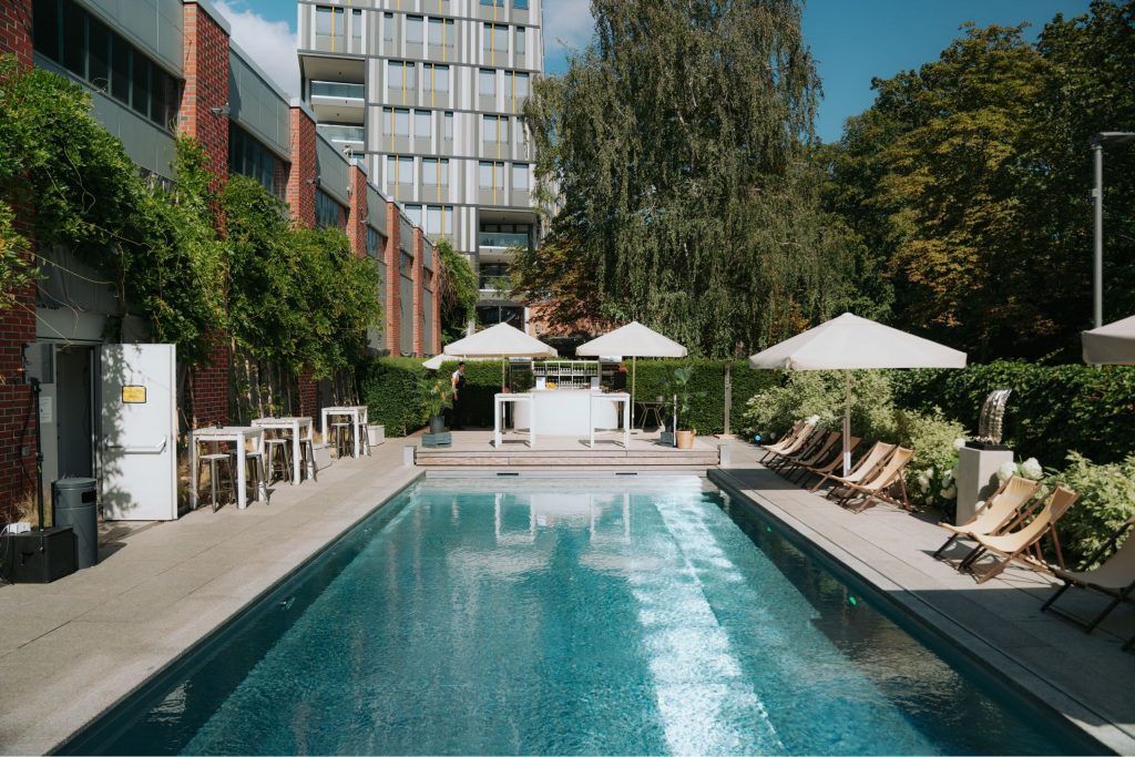 Der EUREF-Pool auf dem Berliner Campus, mit gelben Liegestühlen umrandet, 3 große Sonnenschirme spenden Schatten, im Hintergrund sind Bäume sowie Bürogebäude. An der kurzen Seite des Pools ist im Hintergrund eine Bar aufgebaut