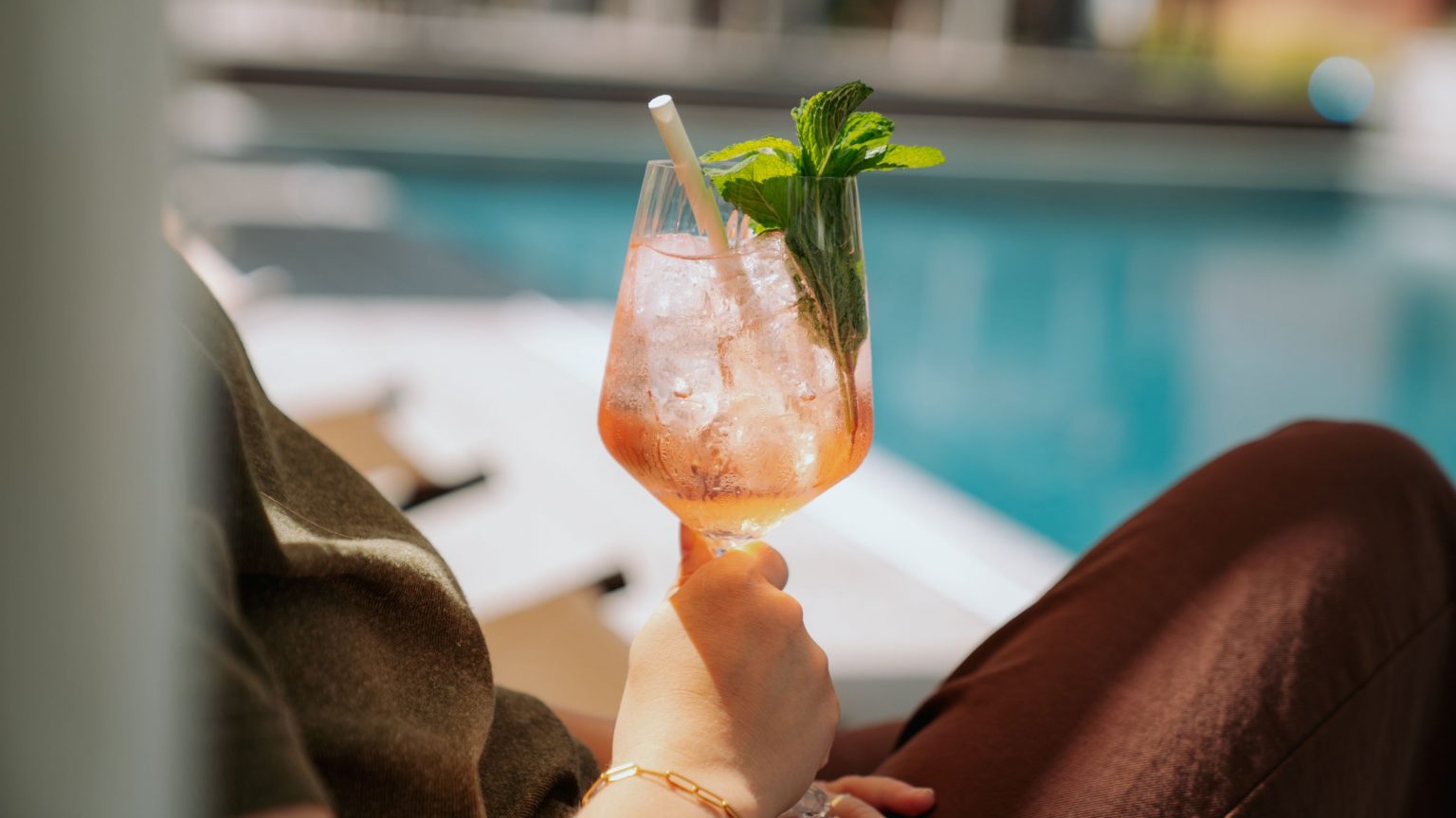 Eine Frau hält einen sommerlichen Cocktain in der Hand, im Hintergrund sieht man den Pool
