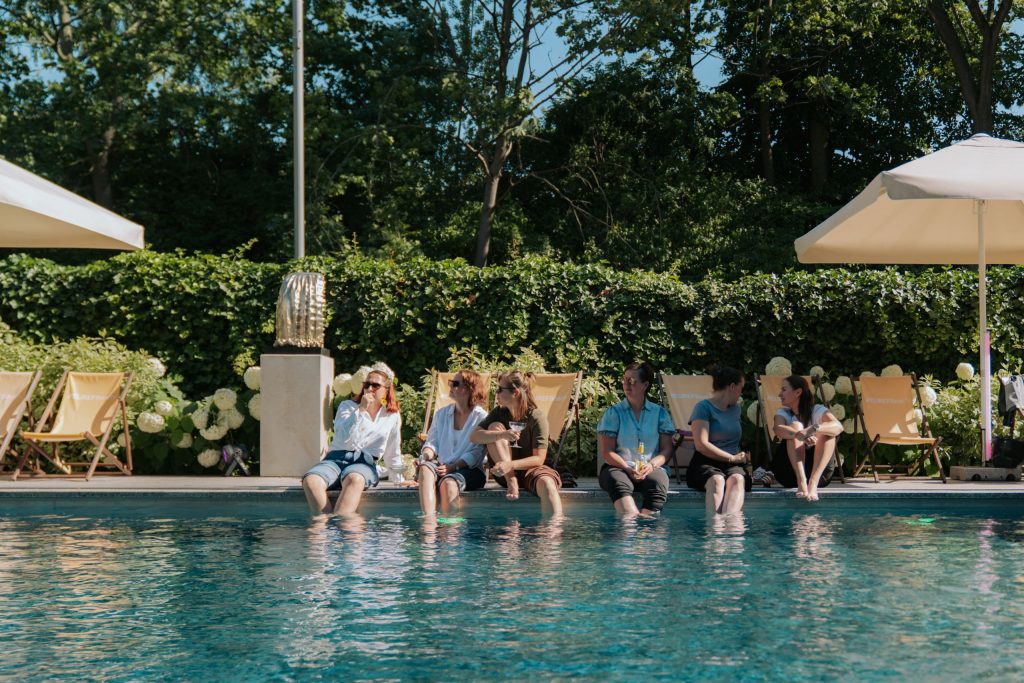 In einer Reihe sitzen Gäste am Pool, die Beine hängen im Wasser, sie unterhalten sich und lachen