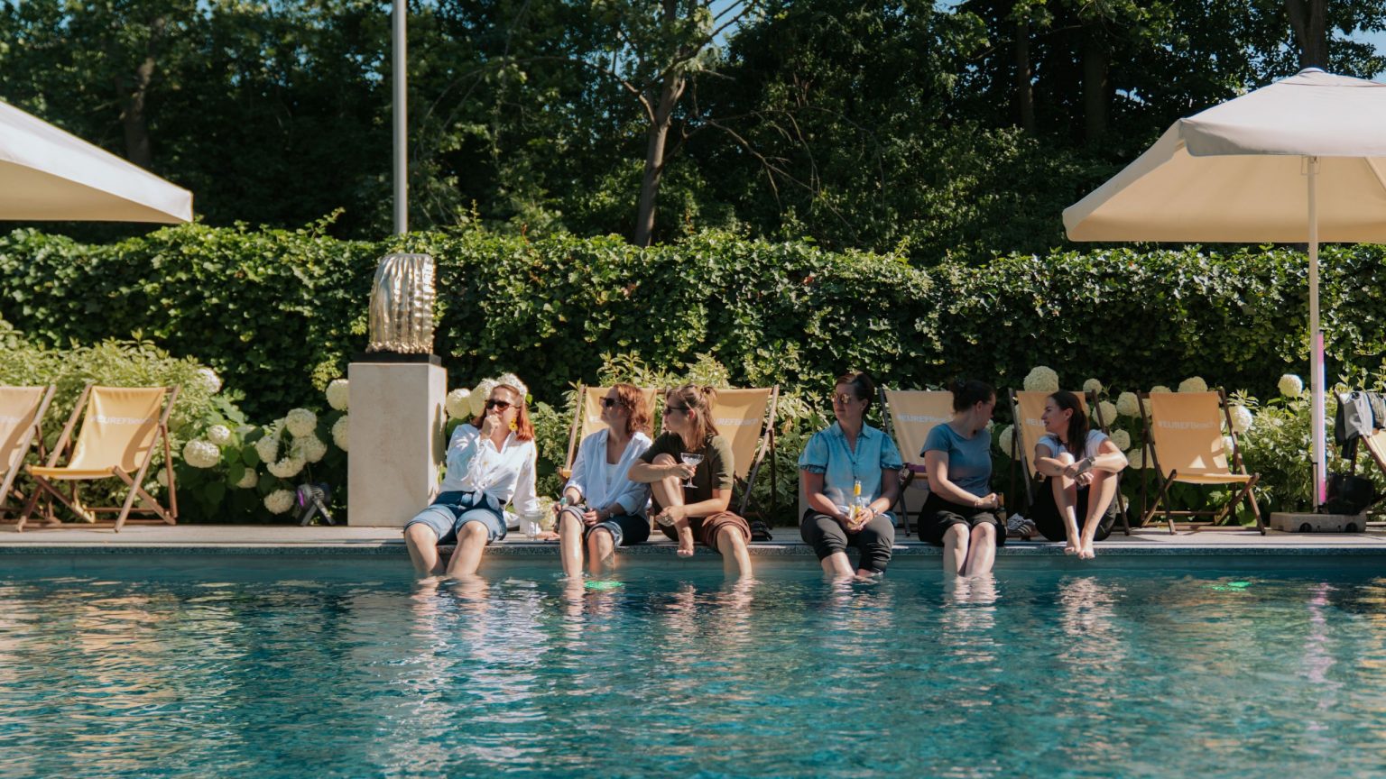 Gäste sitzen am Pool, die Beine hängen im Wasser, sie trinken sommerliche Cocktails