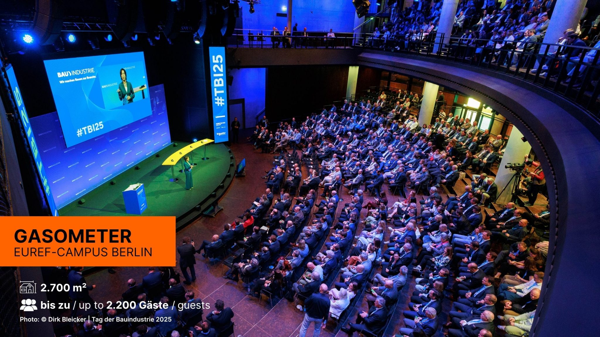 Im Gasometer findet eine Veranstaltung statt, hunderte von Gästen sitzen in Reihen mit Blick auf die große Bühne, eine Rednerin blickt und spricht zum Publikum.