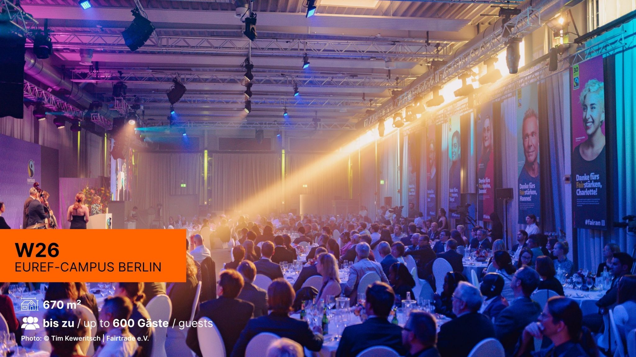 Event in der Location W26 mit stimmungsvollen Licht in blau und lila, Gäste sitzen an runden Tischen bei einer Galaveranstaltung, auf der Bühne spielt eine Live-Band.