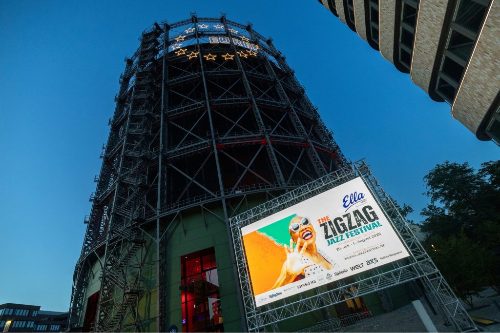 Der Gasometer in Abendstimmung, die Sonne ist bereits untergegangen, auf der LED-Wand vor dem Gasometer ist Werbung für das Jazz Festival zu sehen.
