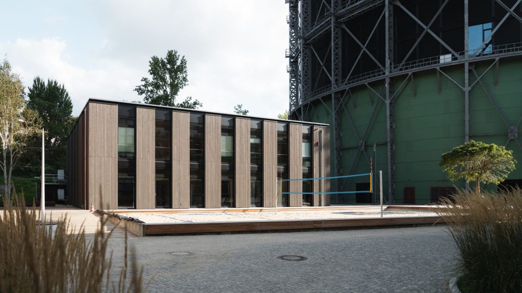 Das Holzhaus mit dem Gasometer im Hintergrund und dem Beachvolleyballfeld
