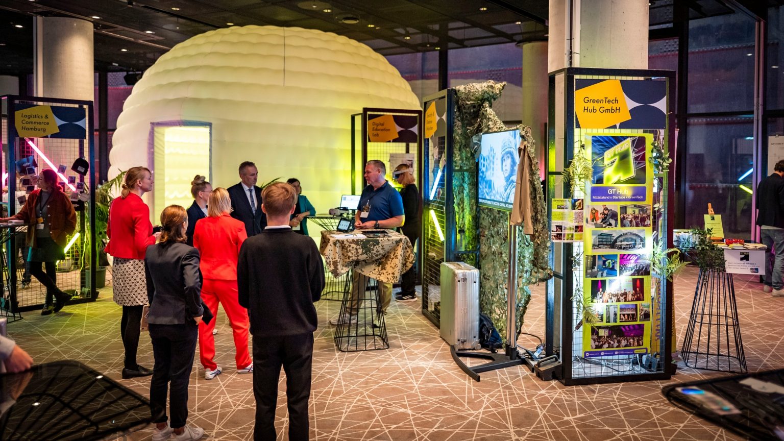 Moderne, im Industrielook gestaltete Ausstellungsflächen mit viel Neonlicht, Gitterwänden, bunten Plakaten, im Hintergrund ein temporäres, aufblasbares Zelt für kleine Vorträge