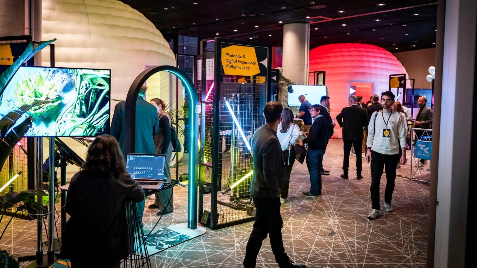 Moderne, im Industrielook gestaltete Ausstellungsflächen mit viel Neonlicht, Gitterwänden, bunten Plakaten, im Hintergrund ein temporäres, aufblasbares Zelt für kleine Vorträge