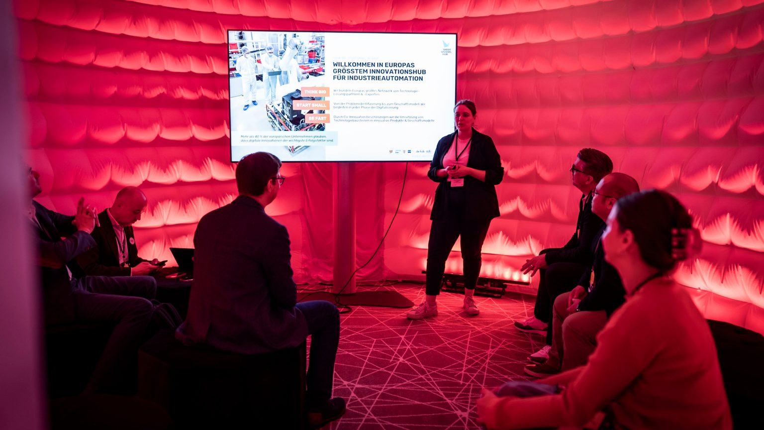 Ein rot leuchtendes temporäres, aufblasbares Zelt für kleine Vorträge, innen sitzen Gäste, eine Rednerin hält neben einem Screen einen Vortrag