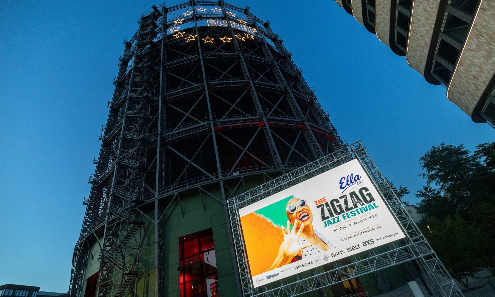 Der Gasometer in Abendstimmung, die Sonne ist bereits untergegangen, auf der LED-Wand vor dem Gasometer ist Werbung für das Jazz Festival zu sehen.