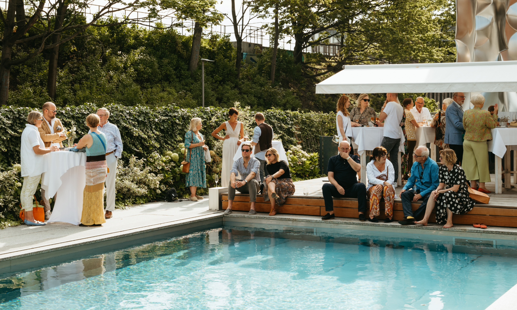 Bei einer Sommerveranstaltung sitzen Gäste an und um einen Pool herum, die Sonne scheint, die Stimmung ist ausgelassen.